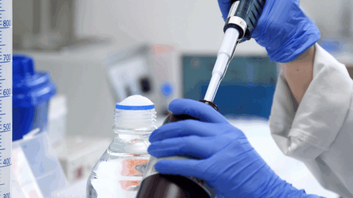 Manos de profesional con guantes azules con pipeta introduciendo líquiso en un frasco en laboratorio